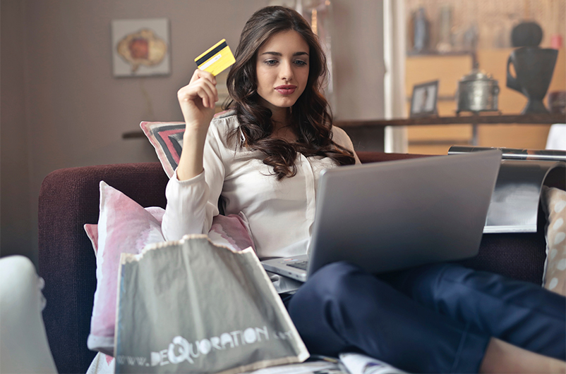 Zdjęcie ukazuje kobietę, która siedzi wygodnie rozparta na kanapie, na kolanach trzymając laptopa. W ręku ma kartę płatniczą, a jej wzrok skierowany jest na ekran. Najprawdopodobniej robi zakupy przez Internet.